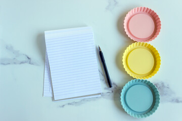 An open paper notebook and pen on a white kitchen table among bright silicone molds. The concept is to create a menu, a list of products and design recipes.