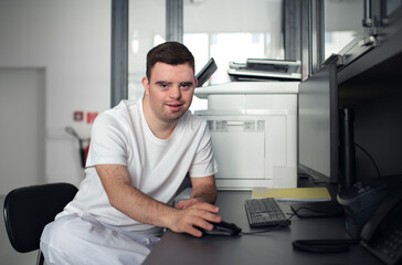 Young man with down syndrome working in hospital office, writing something on computer. Concept of...