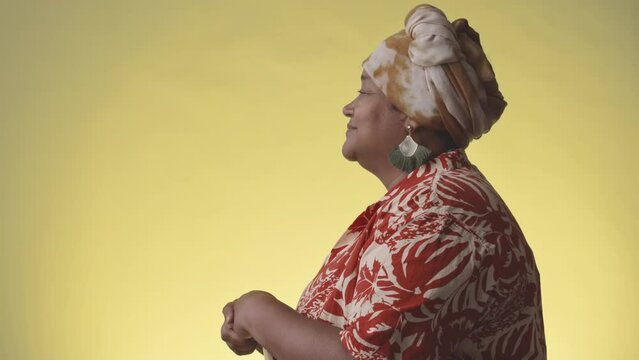 Side view medium portrait of modern African American mature woman in Hawaiian print shirt and turban on her head posing for camera on yellow studio background