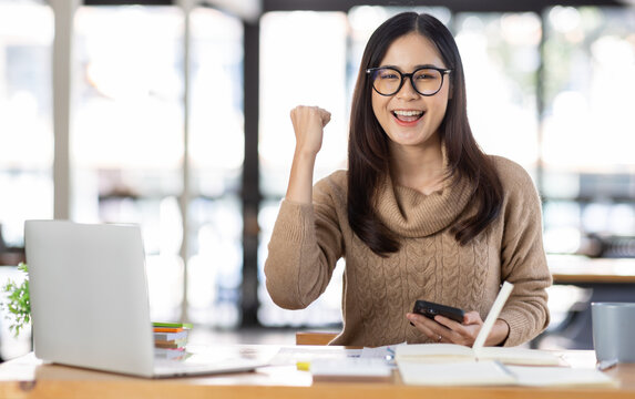 Portrait of Happy Excited young asian woman at workplace office desk, successful Asian female reading good news technology online, employee freelance finance concepts.