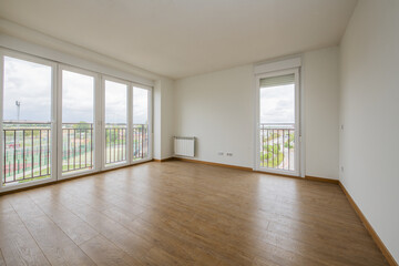 Empty room with light wooden floors, white aluminum radiators, smooth white painted walls and four aluminum and glass doors with views of the city and a second balcony on another wall