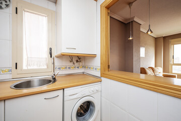 Corner of a small kitchen with white cabinets and light wooden countertops and a serving hatch to the living room
