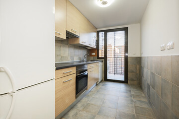 Elongated kitchen with wooden furniture with black countertop, gray tiles and exit to a drying terrace with aluminum and glass doors