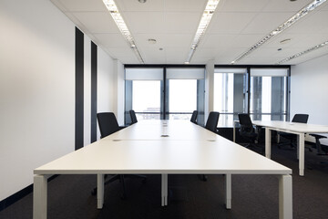 Spacious empty office with brown carpet and long white wooden tables with swivel black seats and technical ceiling and large windows with views