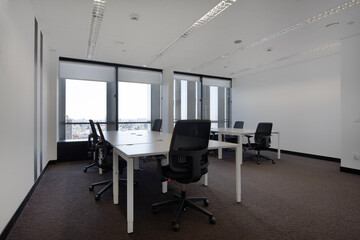 Spacious empty office with brown carpet and white wooden tables with swivel black seats and technical ceiling and large windows with views