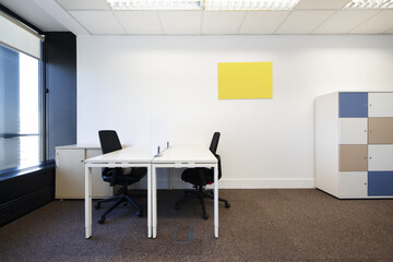 Spacious office with brown carpet, white wooden tables, filing cabinet with colored doors and black swivel chairs and bay window with view
