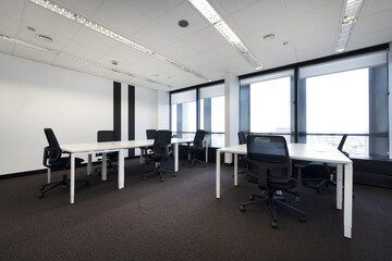 Fototapeta na wymiar Spacious empty office with brown carpet and long white wooden tables with swivel black seats and technical ceiling and large windows with views