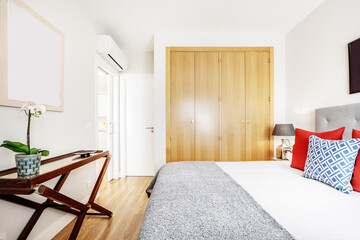Bedroom with wooden furniture with a built-in wardrobe with four doors and a double bed with a headboard upholstered in gray fabric and red and blue cushions