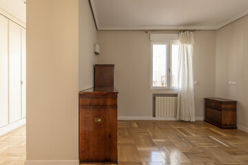 A semi-empty room with some vintage-style wooden furniture with a window with curtains and an exit to a walk-in hallway with Venetian-style white wooden door cabinets