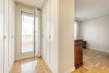 A half-empty room with some old-style wooden furniture, an exit to a corridor with white wooden cabinet doors in Venetian style with access to a terrace through a white aluminum and glass door