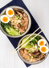 Two bowls of Asian noodle soup, ramen with chicken breast, vegetables, mushrooms, egg and chopsticks on a dark napkin, on a white wooden table. Top view, healthy eating concept