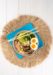 Asian noodle soup, ramen with chicken breast, vegetables, mushrooms, egg and chopsticks on a blue napkin, on a white wooden table. Top view, healthy eating concept