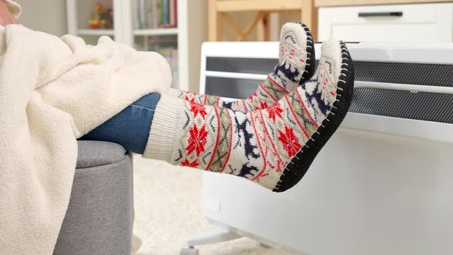 Closeup of woman wearing warm socks sitting at electric heater in living room. Concept of energy crisis, high bills, economy and saving money on monthly utility payments