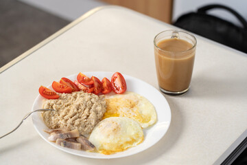 Breakfast of oatmeal with roasted egg, tomato slices, herring slices and a cup of coffee with milk. 