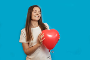 A young beautiful girl smiles happily with her eyes closed and a heart-shaped balloon in her hands on a blue background