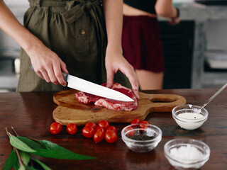 Woman with knife in hand cutting steak meat for frying in kitchen with salt pepper and other spices on table, red cherry tomatoes and herbs, dinner preparation.
