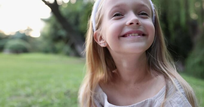 Close up emotional portrait of caucasian smiling teen girl with blonde hair seven years old. Funny cute child in summer park on sunset. Happy kid having fun when looking at camera