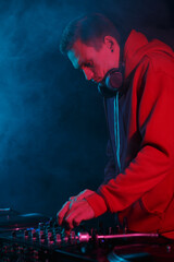 Cool young dj in red hoodie plays music on party in smoke. White male person playing set in night club