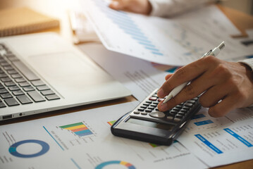 Man accountant use calculator with laptop and holding a pen and taking notes in a notebook and checking financial reports from documents lying on his desk at home office.