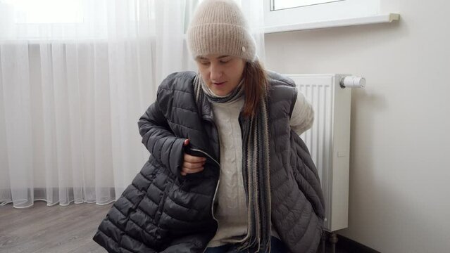 Woman freezing in home putting on warm coat sitting at radiator. Concept of energy crisis, high bills, broken heating system, economy and saving money on monthly utility payments