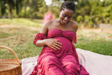 Happy african pregnant woman touching her belly while enjoy a pic nic at city park