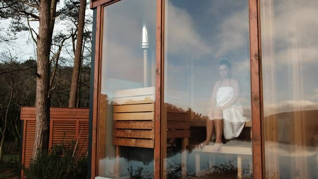 Young woman wrapped in a white towel sitting in a sauna with panoramic window. 