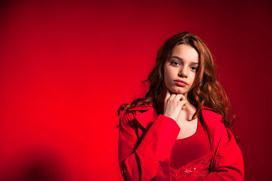 Portrait teenage cover girl with curly hair posing in stylish red coat at red background, looking at camera. Young pretty brunette lady in studio. Fashion style concept. Copy text space for ad