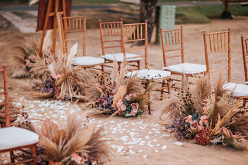 Wedding wooden chairs decorated with flowers. Rustic aisle chairs standing on sand for ceremony on the beach. Natural, shabby, boho wedding decor - 563981984
