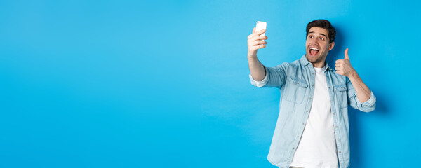 Happy man taking selfie and showing thumb up in approval on blue background, holding mobile phone