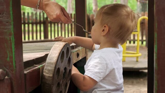 Young mother feeding her baby soon playing on the playground with wooden steering wheel. Kids playing outdoors, children having fun, summer vacation and holiday
