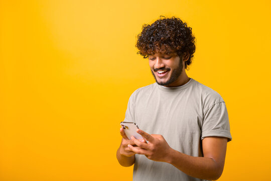 Portrait of cheerful Indian man in casual t-shirt using cellphone and smiling, reading good news message, enjoying mobile application. Curly guy with smartphone in hands