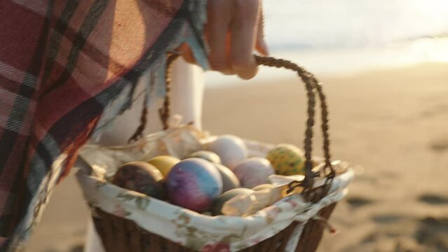 A woman carries a wicker basket with Easter Eggs, The setting sun and the wind blows the plaid on it. Sea sand and waves in the background. Slow motion.
