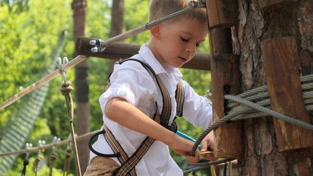 Little boy adjusting safety rope and hook in rope adventure park. Active childhood, healthy lifestyle, kids playing outdoors, children in nature