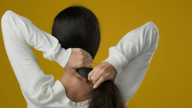 Back view woman with wavy shiny curls hairdo long healthy beautiful hair stylish hairstyle makes ponytail out of her head female Indian ethnic girl model turning to camera in studio yellow background