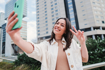 Close up portrait of one young woman smiling taking a selfie portrait with a cellphone, at background city buildings. Front view of a brunette happy girl doing a photo with a smartphone and sharing in