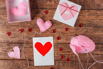 On a wooden background are knitted pink hearts, knitting tools, yarn, a souvenir box and a greeting card. Valentine's day, manual labor, hobby, preparation for the holiday