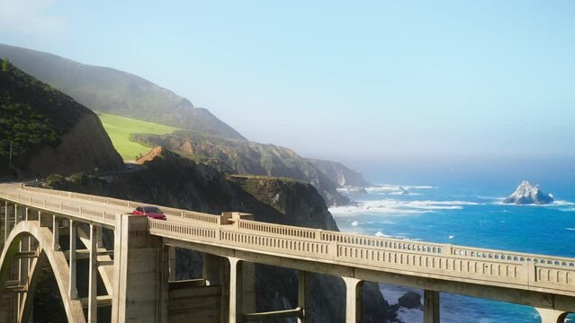 Slow motion electric car driving by arch Bixby bridge. Zero emission vehicle on a scenic road trip by sunny California coast. Cinematic travel route by Pacific Ocean Coast on background RED camera 4K
