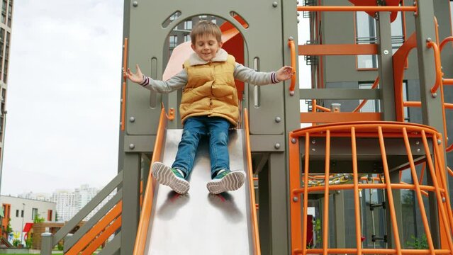 Slow motion of cheerful smiling boy having fun sliding down the slide at new playground. Active child, sports and development, kids playing outdoors.