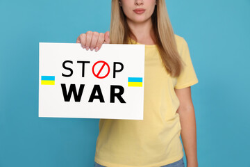 Woman holding poster with words Stop War and Ukrainian flags on light blue background, closeup