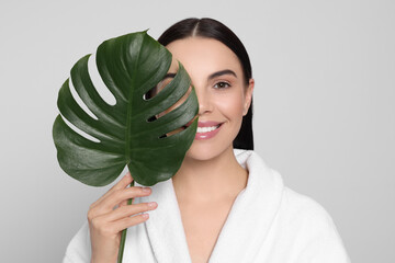 Woman in bathrobe holding leaf of monstera on light grey background. Spa treatment