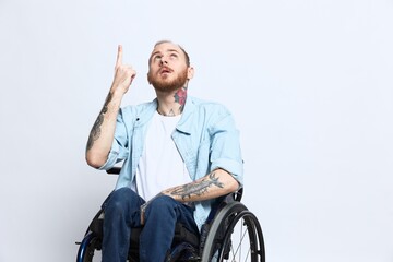 A man in a wheelchair and pointing a finger at, copy space, with tattoos on his hands sits on a gray studio background, health concept a person with disabilities, a real person