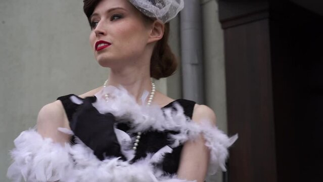 A woman stands in a white feather boa on the porch against a light wall. Slow motion of a woman in a historical costume of the 20-30s of the XX century. High quality FullHD footage