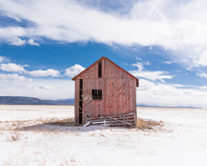 Red abandoned barn in an open field, San Luis Valley, Colorado, with fence, bright blue sky, sunshine, snow, and clouds