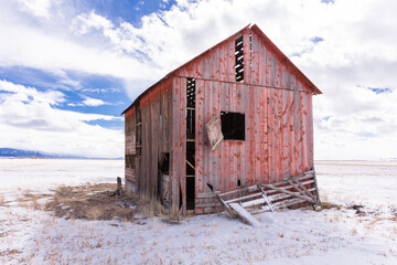 Red abandoned barn in an open field, San Luis Valley, Colorado, with fence, bright blue sky, sunshine, snow, and clouds