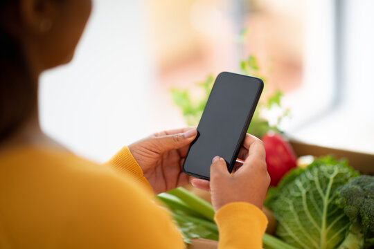 Young Black Woman Typing On Smartphone With Blank Screen Over Package With Groceries, Organic Vegetables In Kitchen