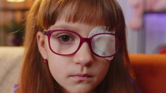 Sick ill toddler girl face looking at camera having protective bandage plaster patch scratch on one eye. Injury trauma blind. Child kid wearing glasses at home in room. Medical eyesight vision therapy