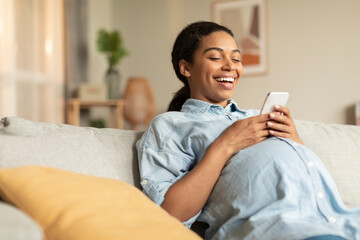 Joyful black pregnant woman using smartphone while relaxing on couch, watching funny videos and laughing, copy space