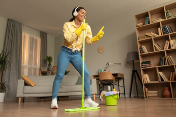Playful african american lady in headphones making cleaning at home, singing and using mop as...