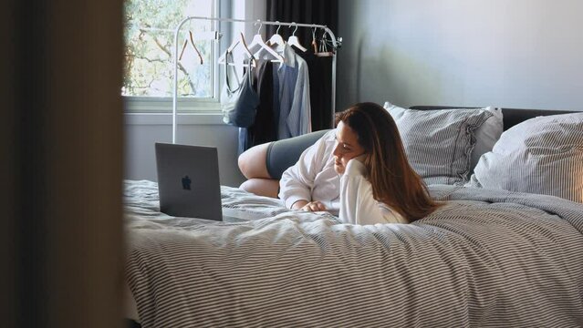 Calm overweight woman laying at the bed and looking at laptop while relaxing