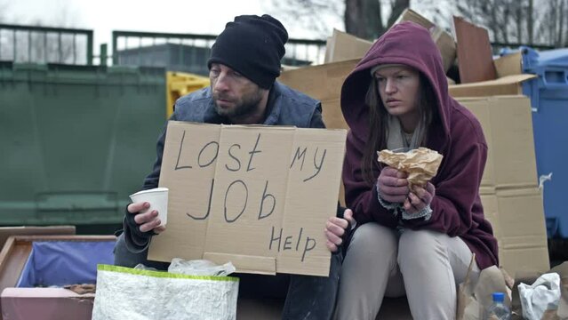 Two dirty and poorly dressed homeless people, a man and a woman, sit by a pile of rubbish with a handwritten LAST MY JOB HELP poster. Passer-by tosses a few coins into a begging cup.